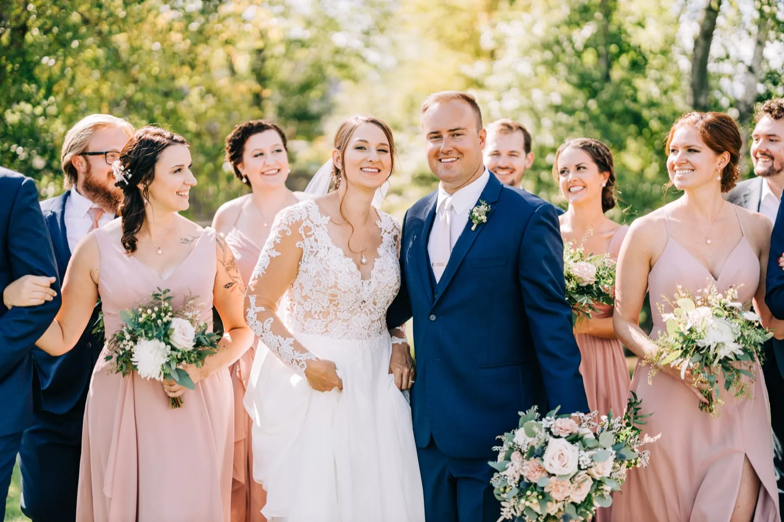 The bride and groom stand smiling at the center of their bridal party, with bridesmaids in blush dresses holding bouquets and groomsmen in navy suits flanking them on a sunlit lawn. — Tim Larsen Photography, Brainerd Lakes MN