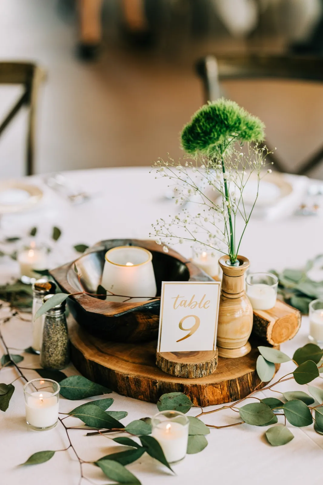 A reception centerpiece featuring a wooden bowl, candle, table number nine card, and a green dianthus stem in a wooden vase atop a wood disc made by the bride's family, surrounded by trailing eucalyptus. — Tim Larsen Photography, Brainerd Lakes MN