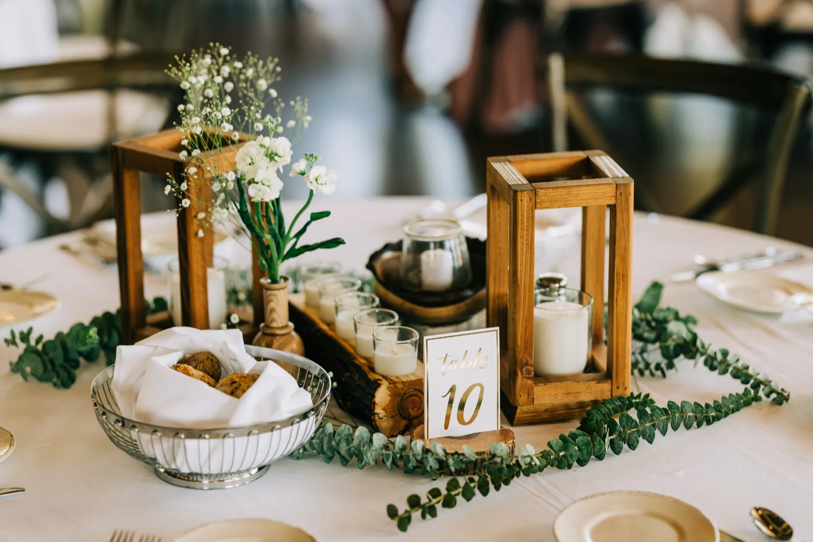 A reception centerpiece featuring wooden lanterns with pillar candles, a bread basket, baby's breath florals, and a gold-lettered table number 10 card resting on eucalyptus greenery. — Tim Larsen Photography, Brainerd Lakes MN