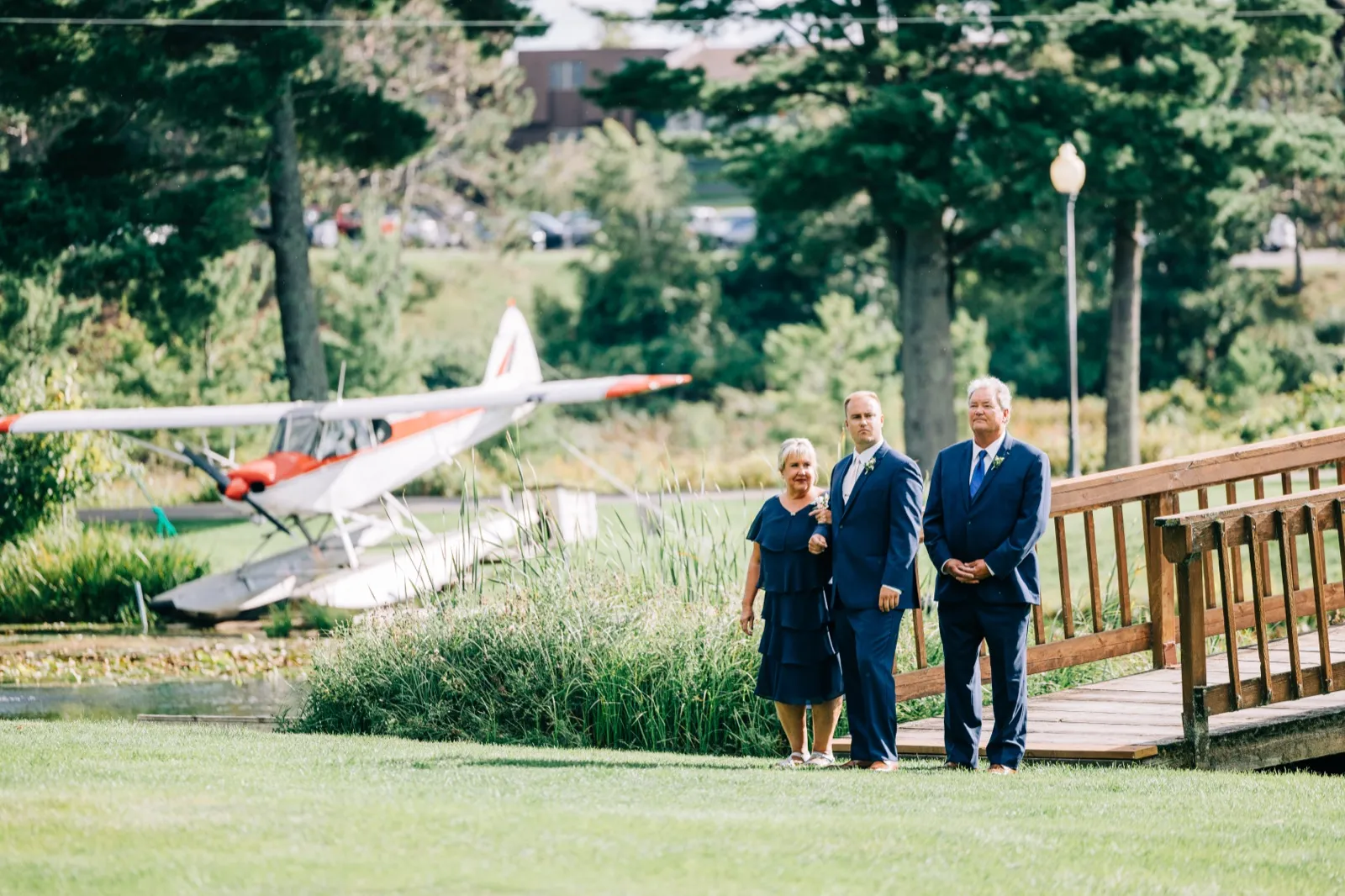 The groom stands between his parents on a grassy lawn at Madden's on Gull Lake, with a vintage red and white floatplane parked behind them and a wooden footbridge to their right. — Tim Larsen Photography, Brainerd Lakes MN