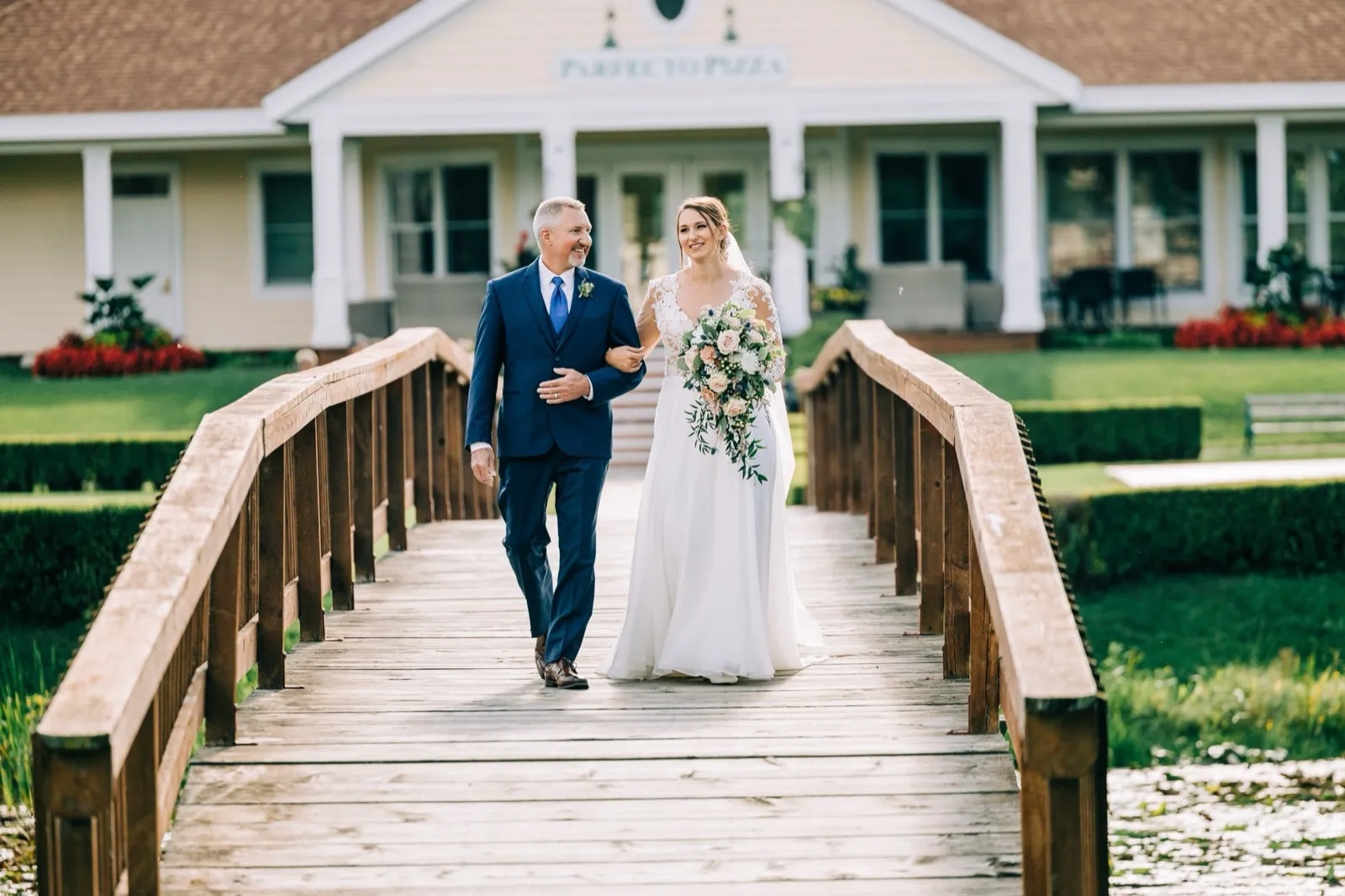 The bride walks across a wooden arched footbridge with her father, holding a cascading bouquet, with the Madden's lodge building behind them. — Tim Larsen Photography, Brainerd Lakes MN
