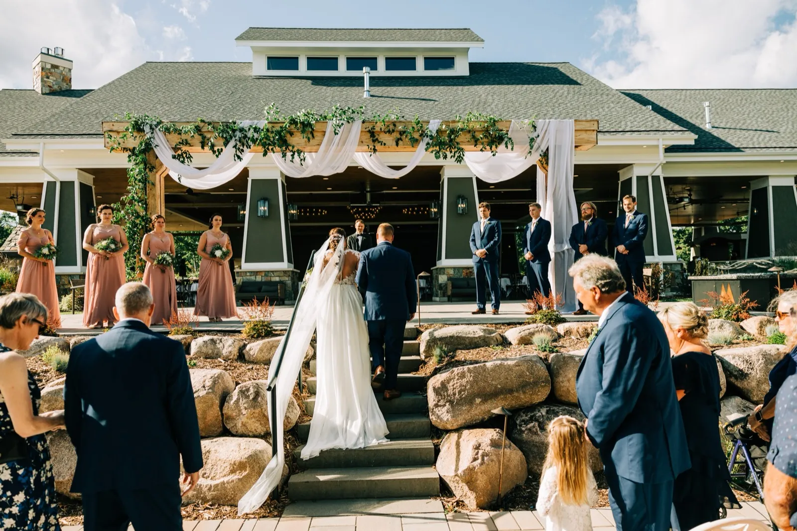 The bride climbs stone steps toward the draped ceremony pavilion at Madden's on Gull Lake while bridesmaids, groomsmen, and guests watch from either side. — Tim Larsen Photography, Brainerd Lakes MN