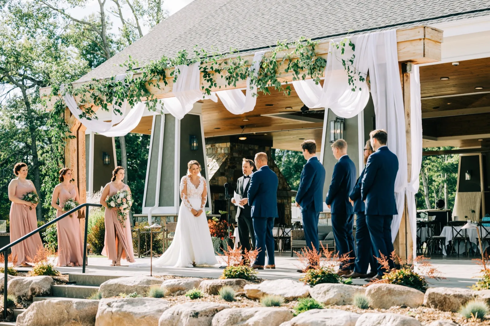 The bride and groom stand under a draped pavilion arch covered in greenery while the officiant reads, with the bridal party lined up on either side at an outdoor ceremony at Madden's on Gull Lake. — Tim Larsen Photography, Brainerd Lakes MN