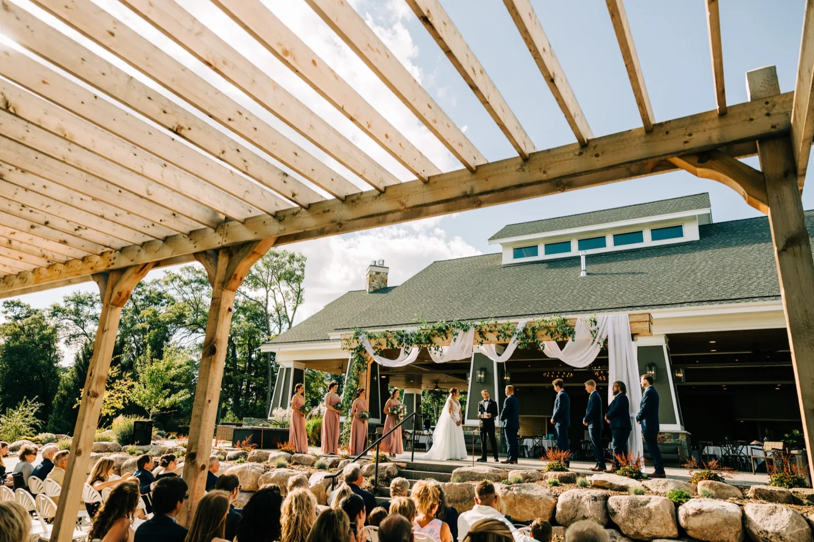 A wide view of an outdoor ceremony at Madden's on Gull Lake, with the bride, groom, bridal party, and officiant on a covered stage framed by the wooden beams of a pergola, while seated guests watch from the foreground. — Tim Larsen Photography, Brainerd Lakes MN