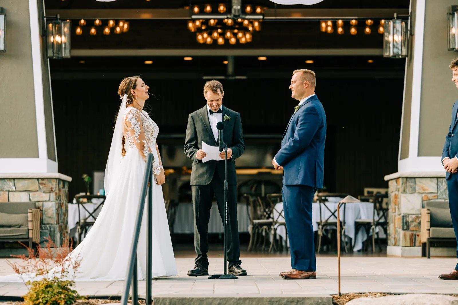The bride laughs head-tilted-back as the officiant reads from a paper at the microphone and the groom watches her, all standing under a chandelier-lit pavilion at an outdoor ceremony. — Tim Larsen Photography, Brainerd Lakes MN