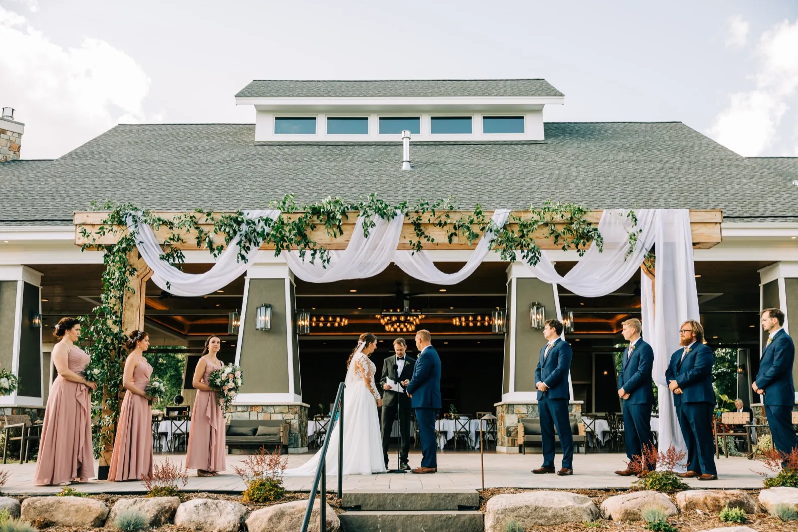 A wide symmetrical view of the bride and groom exchanging vows beneath a greenery-and-drapery pavilion at Madden's on Gull Lake, flanked by their bridal party. — Tim Larsen Photography, Brainerd Lakes MN