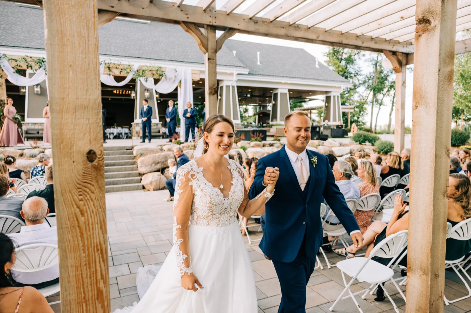 The bride and groom walk hand-in-hand down the aisle after their ceremony, smiling broadly as guests watch from white folding chairs beneath a wooden pergola. — Tim Larsen Photography, Brainerd Lakes MN