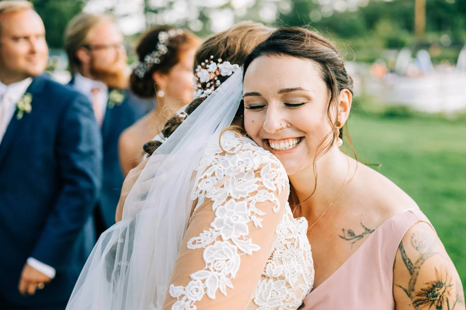 A guest in a pink dress closes her eyes and smiles broadly while embracing the bride, whose lace sleeve and veil fill the foreground. — Tim Larsen Photography, Brainerd Lakes MN