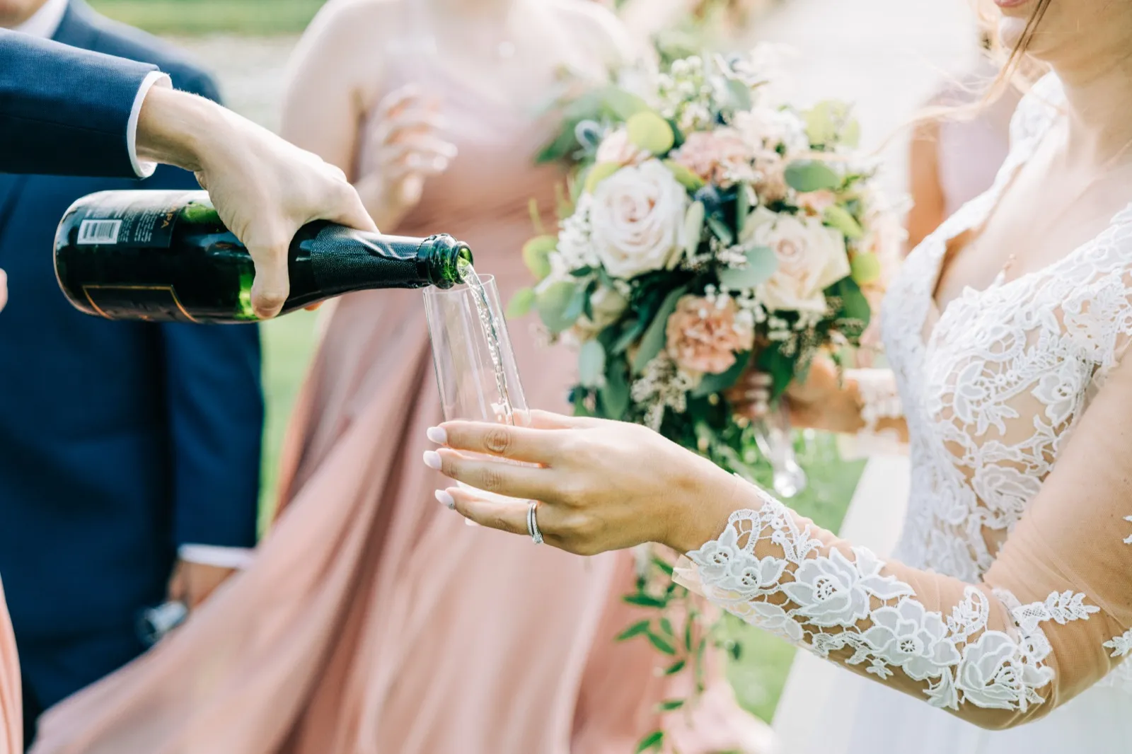 The groom pours champagne into a flute held by the bride, whose lace-sleeved arm and pastel bouquet fill the frame alongside a bridesmaid in a blush dress. — Tim Larsen Photography, Brainerd Lakes MN