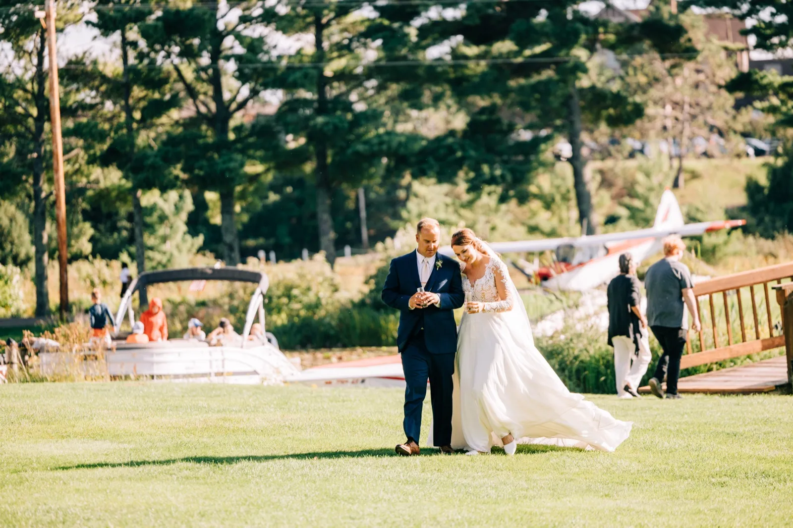 The bride and groom walk together across a sunlit lawn at Madden's on Gull Lake with a seaplane and pontoon boat visible behind them. — Tim Larsen Photography, Brainerd Lakes MN