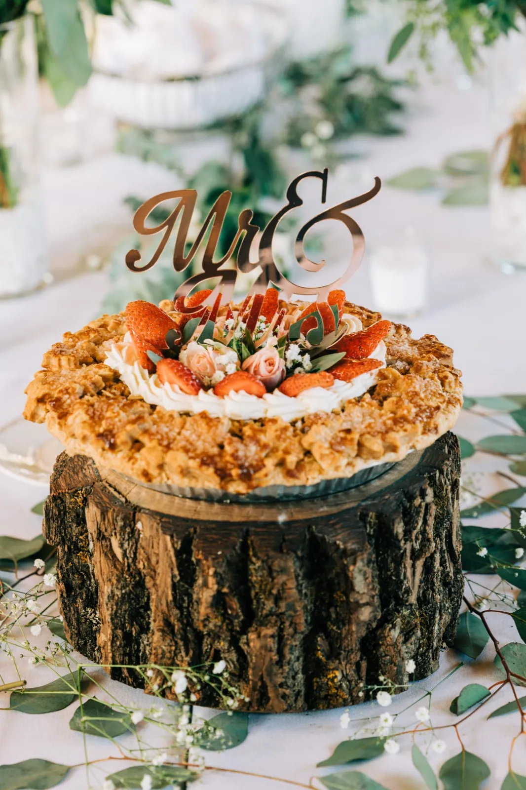 A strawberry-topped wedding pie with a 'Mr & Mrs' topper sits on a bark log slab surrounded by eucalyptus and baby's breath. — Tim Larsen Photography, Brainerd Lakes MN