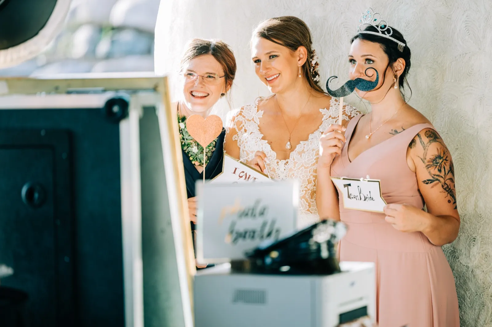 The bride and two bridesmaids smile and laugh while holding photo booth props including a heart, a glittery mustache, and a Team Bride sign. — Tim Larsen Photography, Brainerd Lakes MN