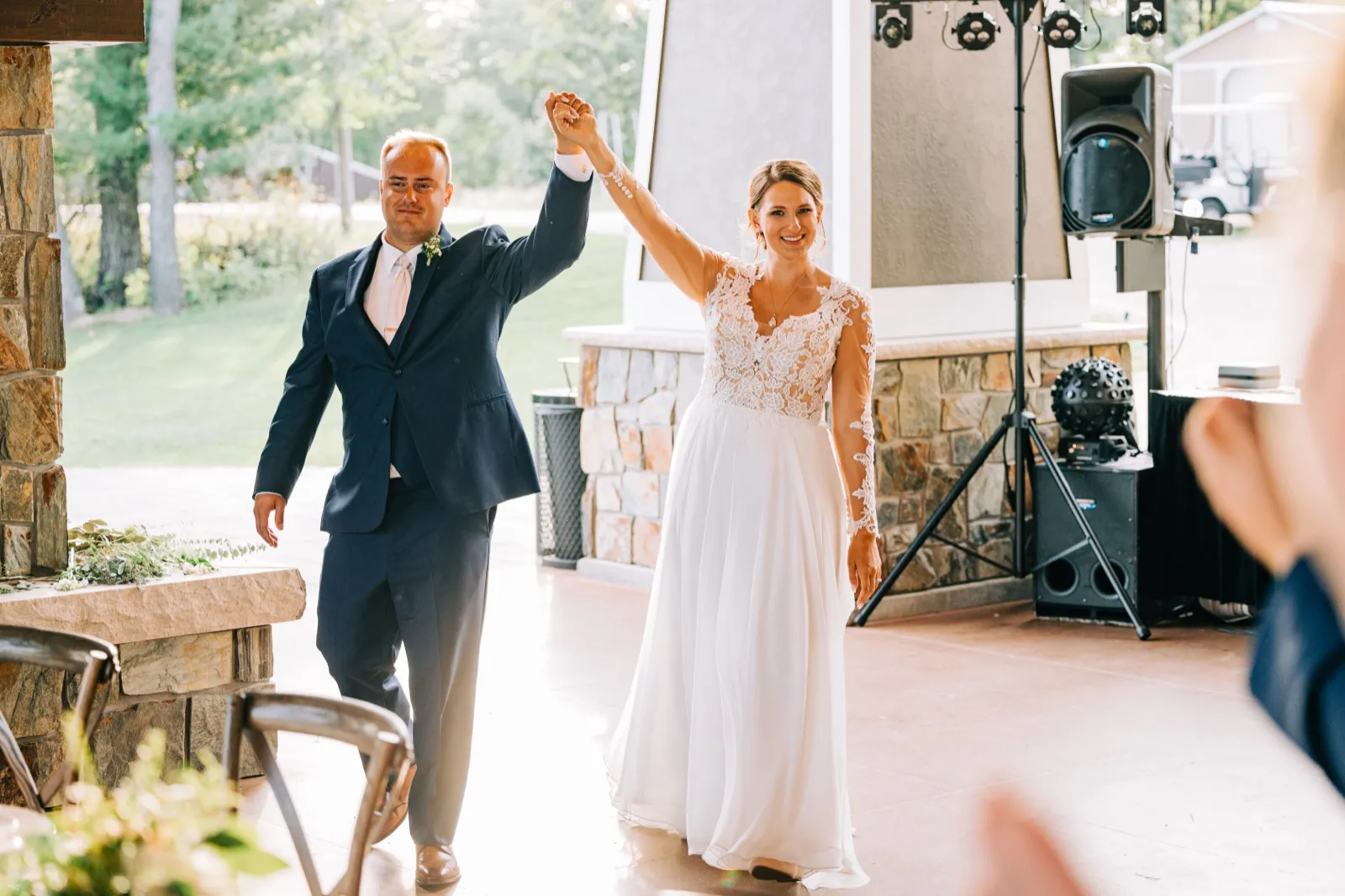 The bride and groom raise their joined hands overhead as they enter the reception pavilion past a stone fireplace and DJ setup. — Tim Larsen Photography, Brainerd Lakes MN