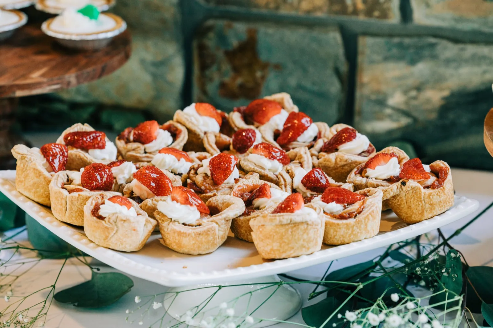 A platter of mini strawberry tarts topped with whipped cream sits on a white pedestal stand in front of a stone fireplace. — Tim Larsen Photography, Brainerd Lakes MN