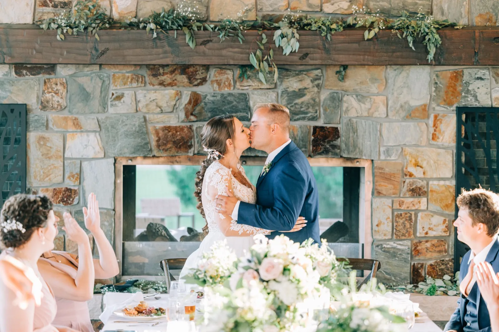 The bride and groom kiss at their head table in front of a massive stone fireplace draped in greenery while bridal party members clap on either side. — Tim Larsen Photography, Brainerd Lakes MN