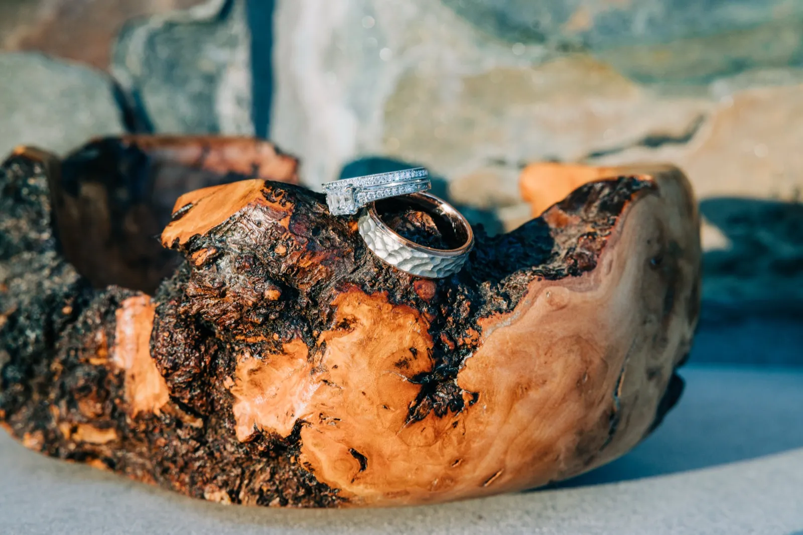 A diamond engagement ring and a hammered silver wedding band rest together in a wooden bowl carved by the bride's grandfather, against a blue-grey stone background. — Tim Larsen Photography, Brainerd Lakes MN