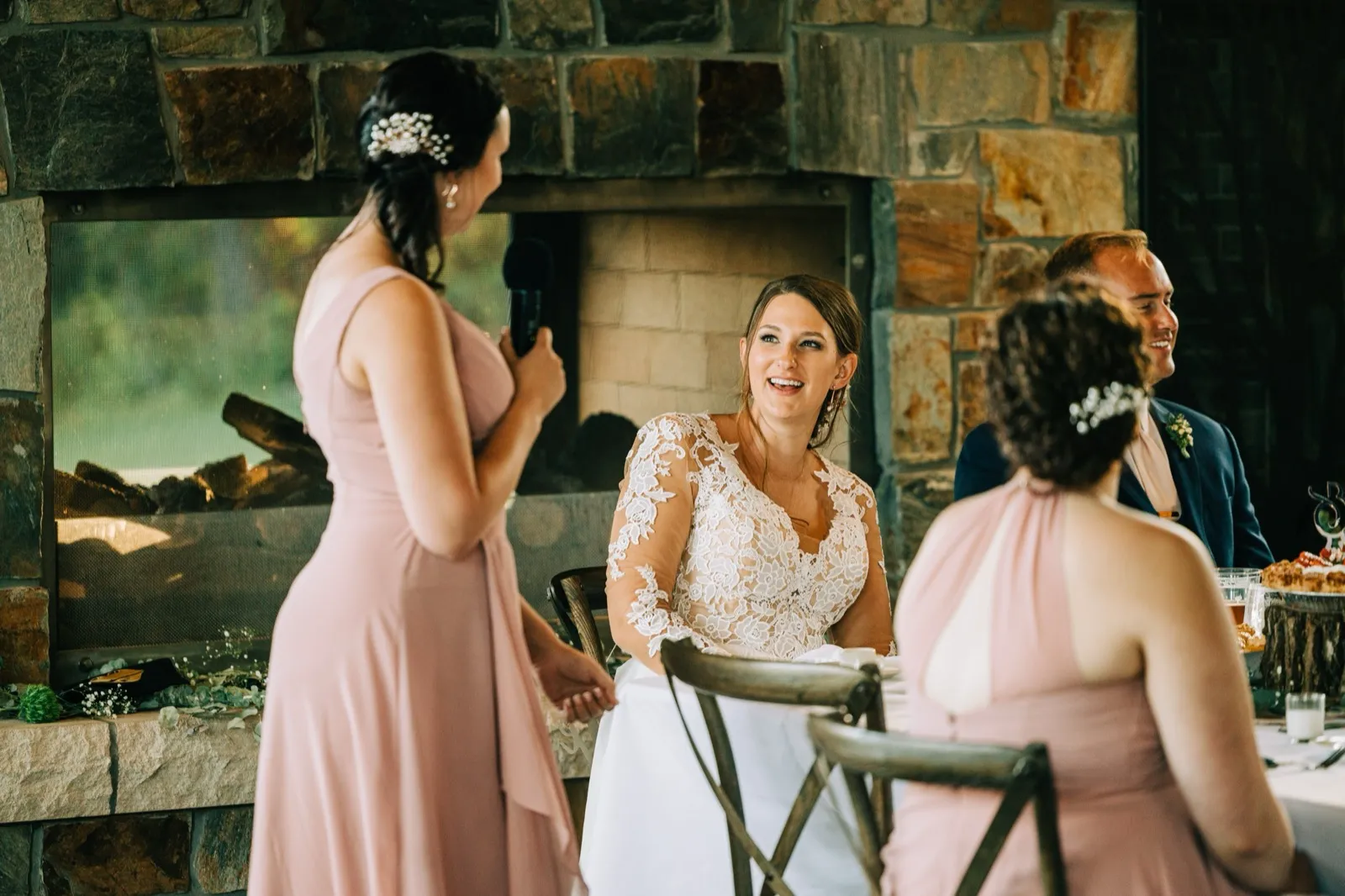 The bride laughs in a lace gown while a bridesmaid in pink holds a microphone during a toast in front of a large stone fireplace. — Tim Larsen Photography, Brainerd Lakes MN