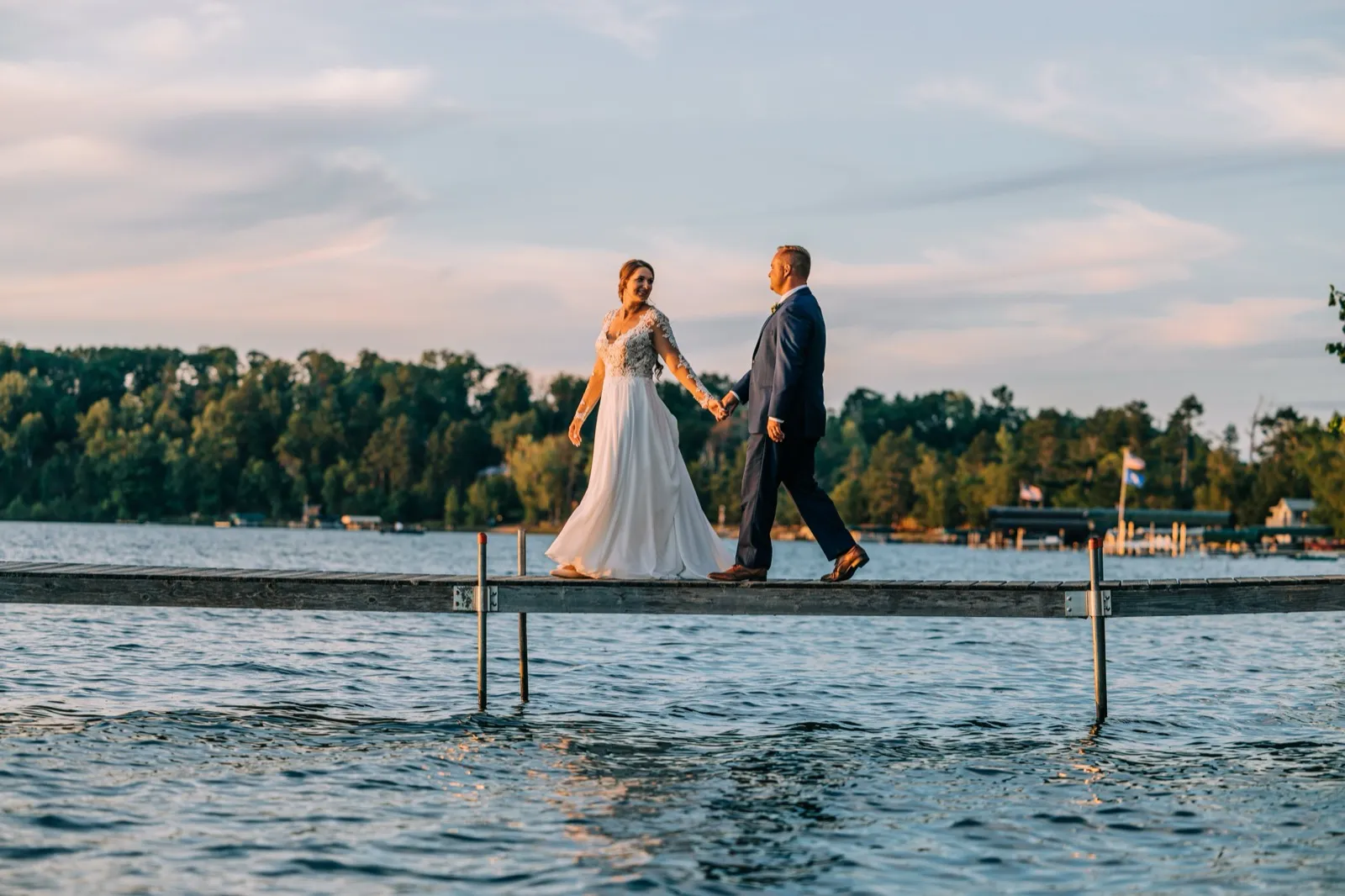 The bride and groom walk hand in hand across a long wooden dock extending over Gull Lake at sunset, with a forested shoreline behind them. — Tim Larsen Photography, Brainerd Lakes MN