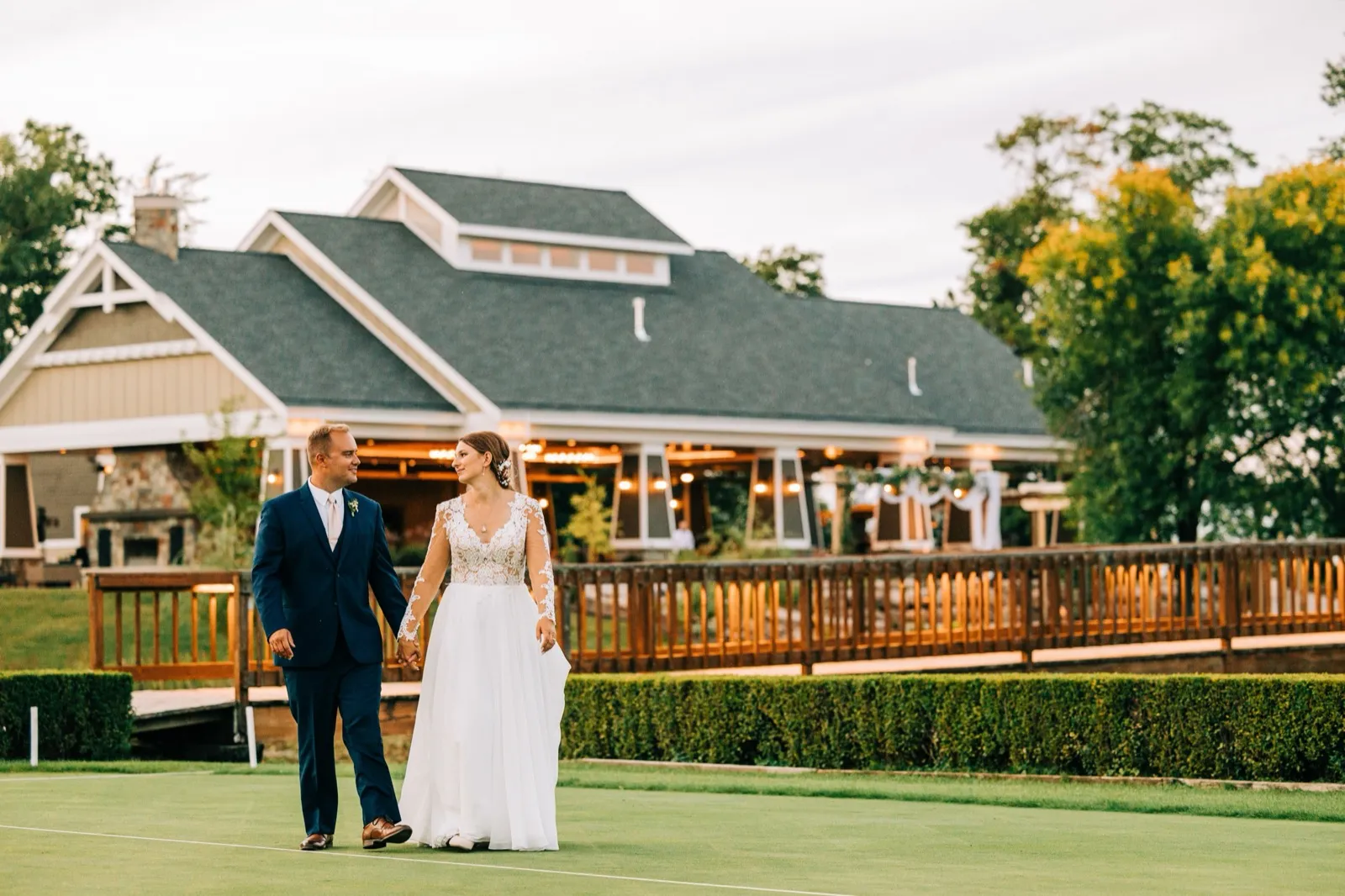 The bride and groom walk hand-in-hand across a lawn at dusk with the warmly lit pavilion of Madden's on Gull Lake glowing behind them. — Tim Larsen Photography, Brainerd Lakes MN