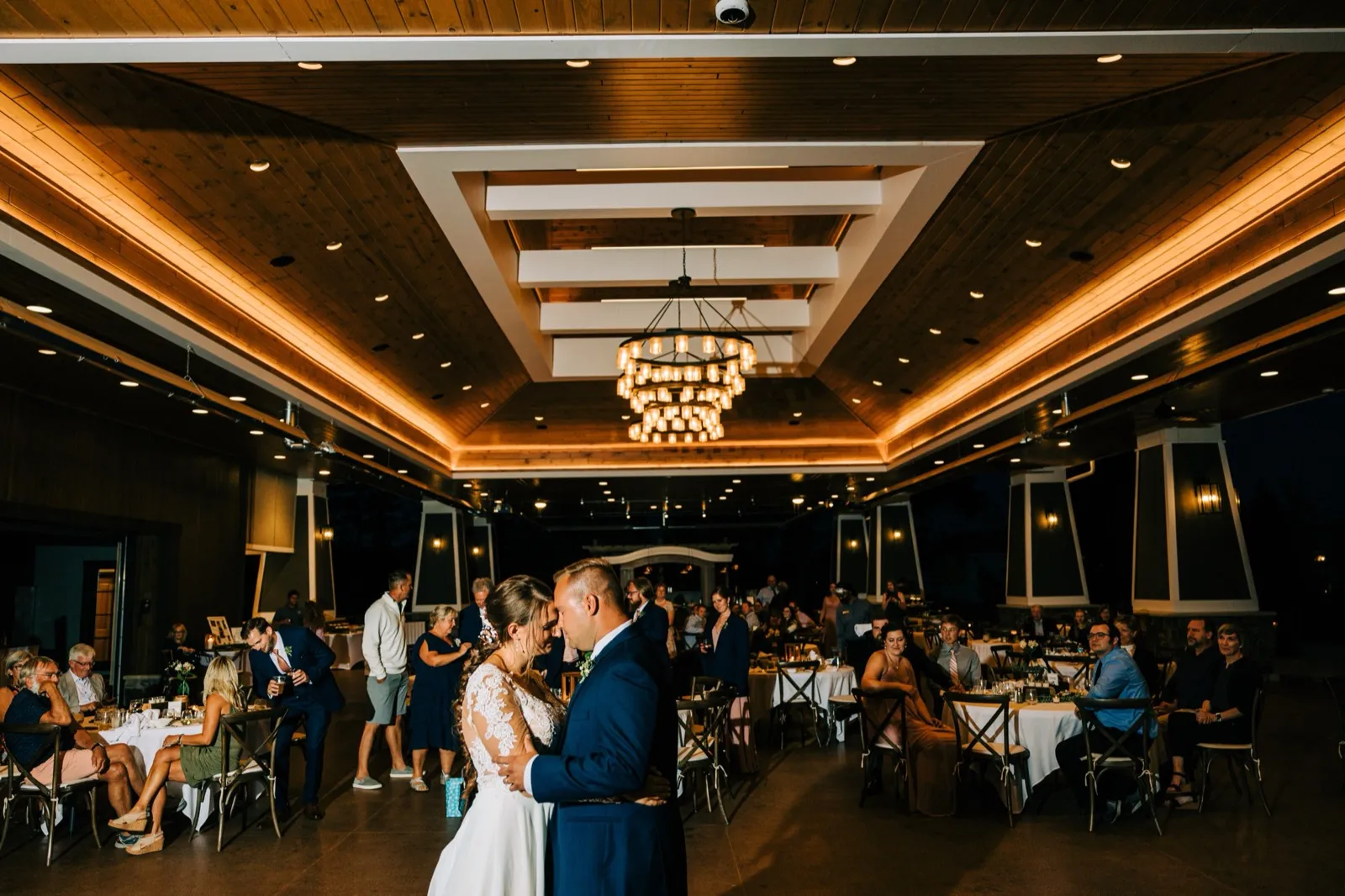 The couple shares a slow dance at the center of a warmly lit pavilion as seated guests watch beneath a tiered chandelier and exposed wood beams. — Tim Larsen Photography, Brainerd Lakes MN