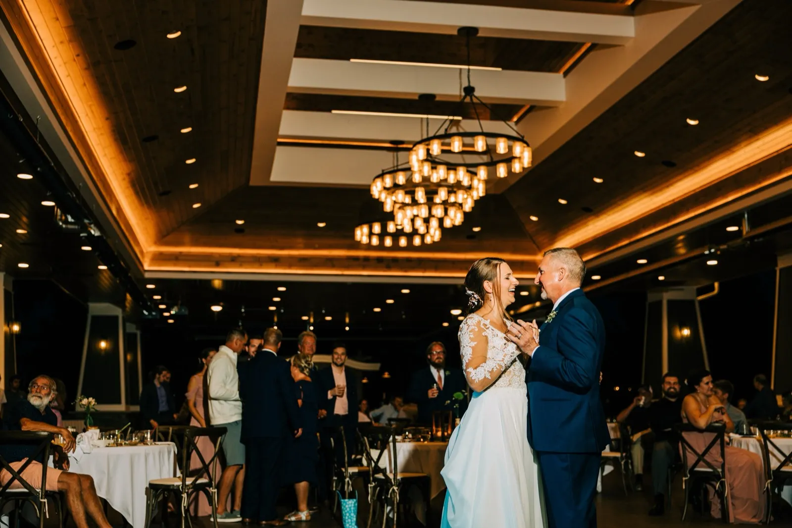 The bride and groom dance and laugh together on a reception dance floor beneath tiered circular chandeliers in a warmly lit wood-ceilinged ballroom while seated guests watch. — Tim Larsen Photography, Brainerd Lakes MN