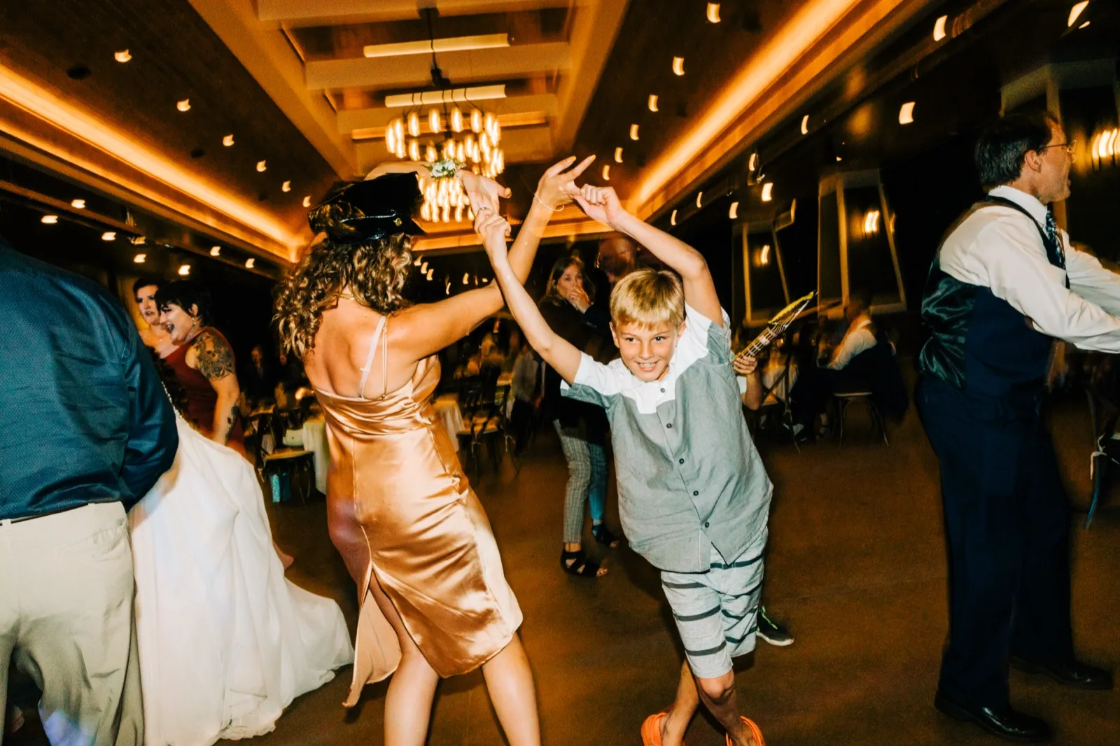 A young boy throws his arms up grinning at the camera while a bridesmaid in a copper satin dress spins beside him on a chandelier-lit dance floor. — Tim Larsen Photography, Brainerd Lakes MN