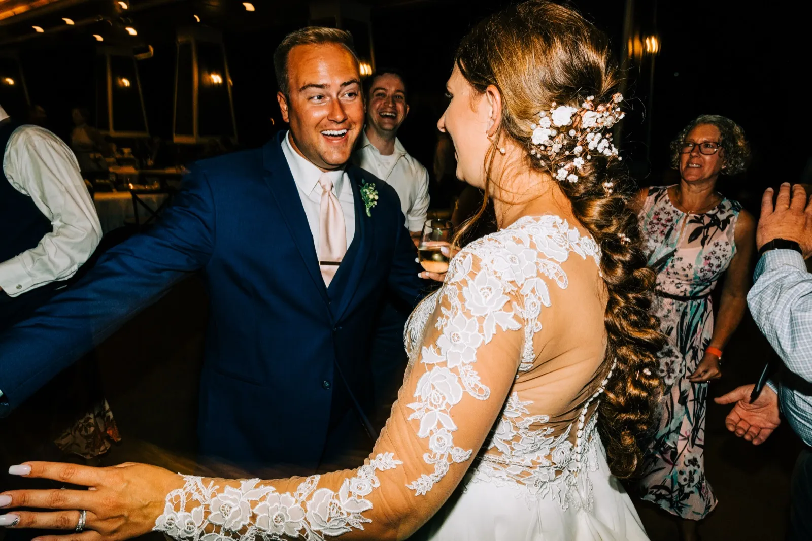 The groom laughs with wide-eyed delight as he dances toward the bride, whose lace-sleeved back and floral-pinned braid fill the foreground. — Tim Larsen Photography, Brainerd Lakes MN