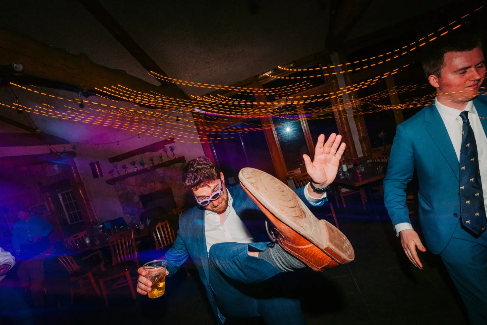 A guest in sunglasses holding a beer kicks his leg high toward the camera under streaks of string lights while a groomsman walks past on the right — Tim Larsen Photography, Brainerd Lakes MN