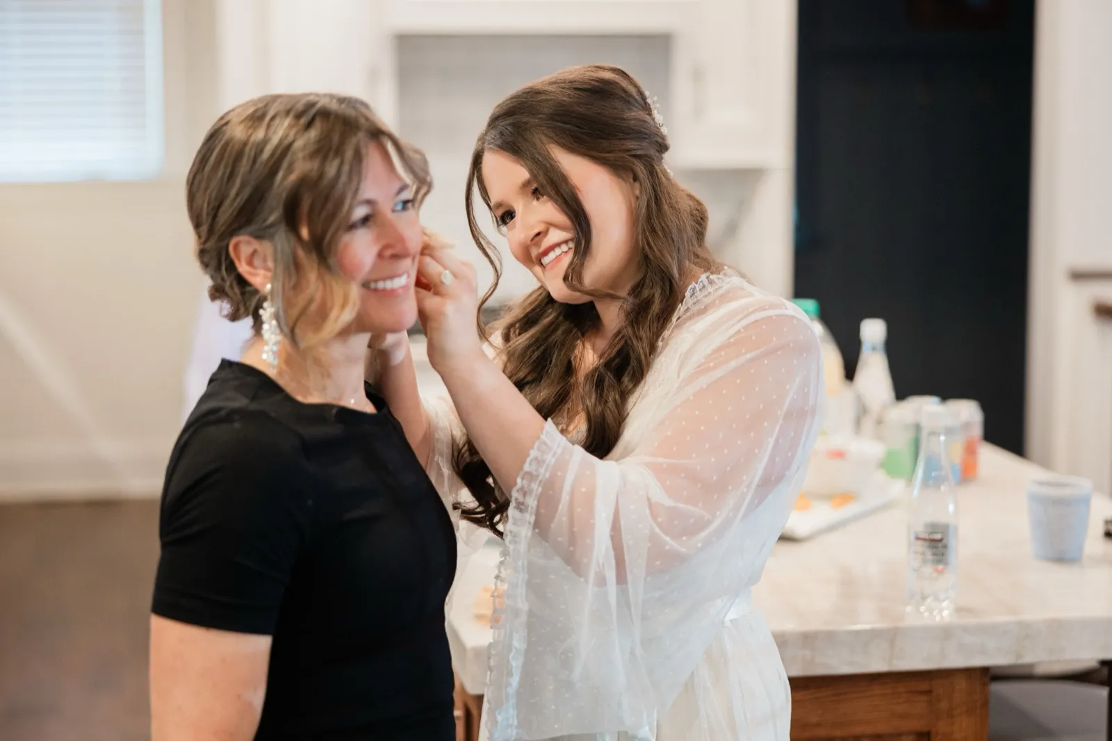 The bride, in her lace robe, holds her mother's face as the two laugh together in the getting-ready room — Tim Larsen Photography, Brainerd Lakes MN