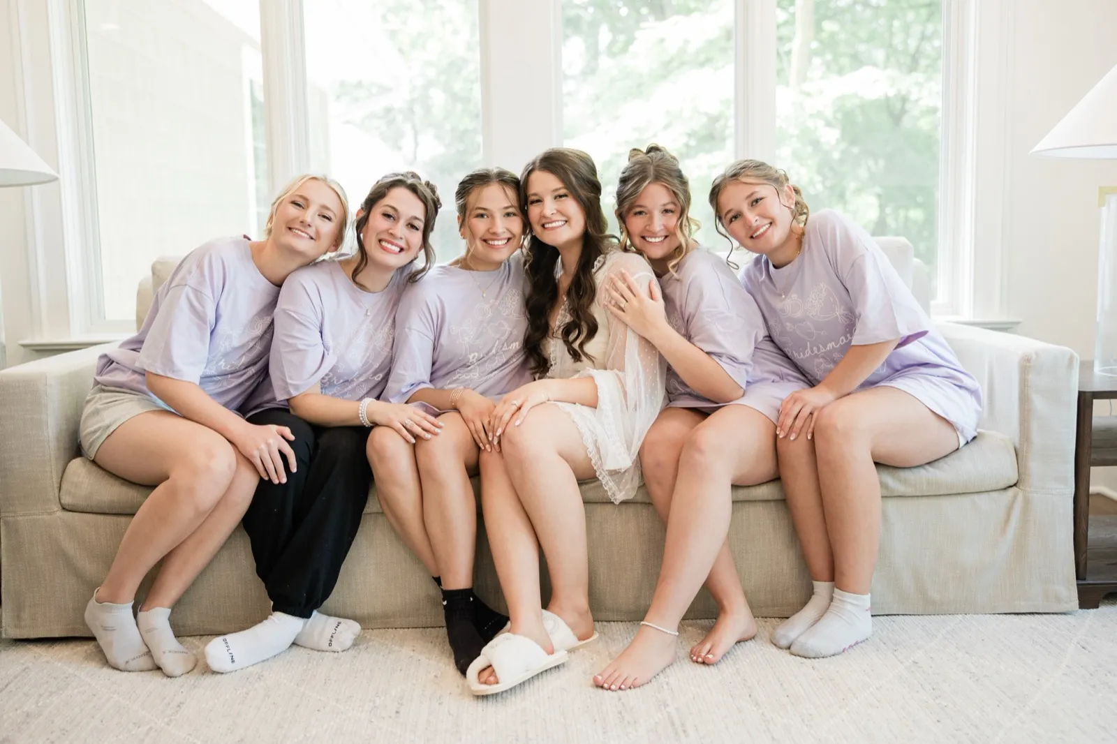 The bride, in a lace robe, sits centered on a sofa among five bridesmaids in matching lavender shirts, all smiling at the camera — Tim Larsen Photography, Brainerd Lakes MN