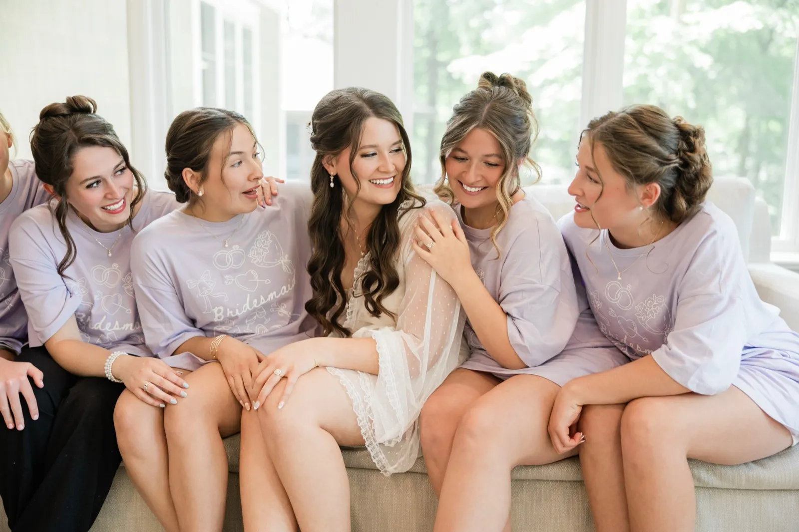 The bride laughs with her head tilted as her bridesmaids lean in around her on the sofa, mid-conversation — Tim Larsen Photography, Brainerd Lakes MN