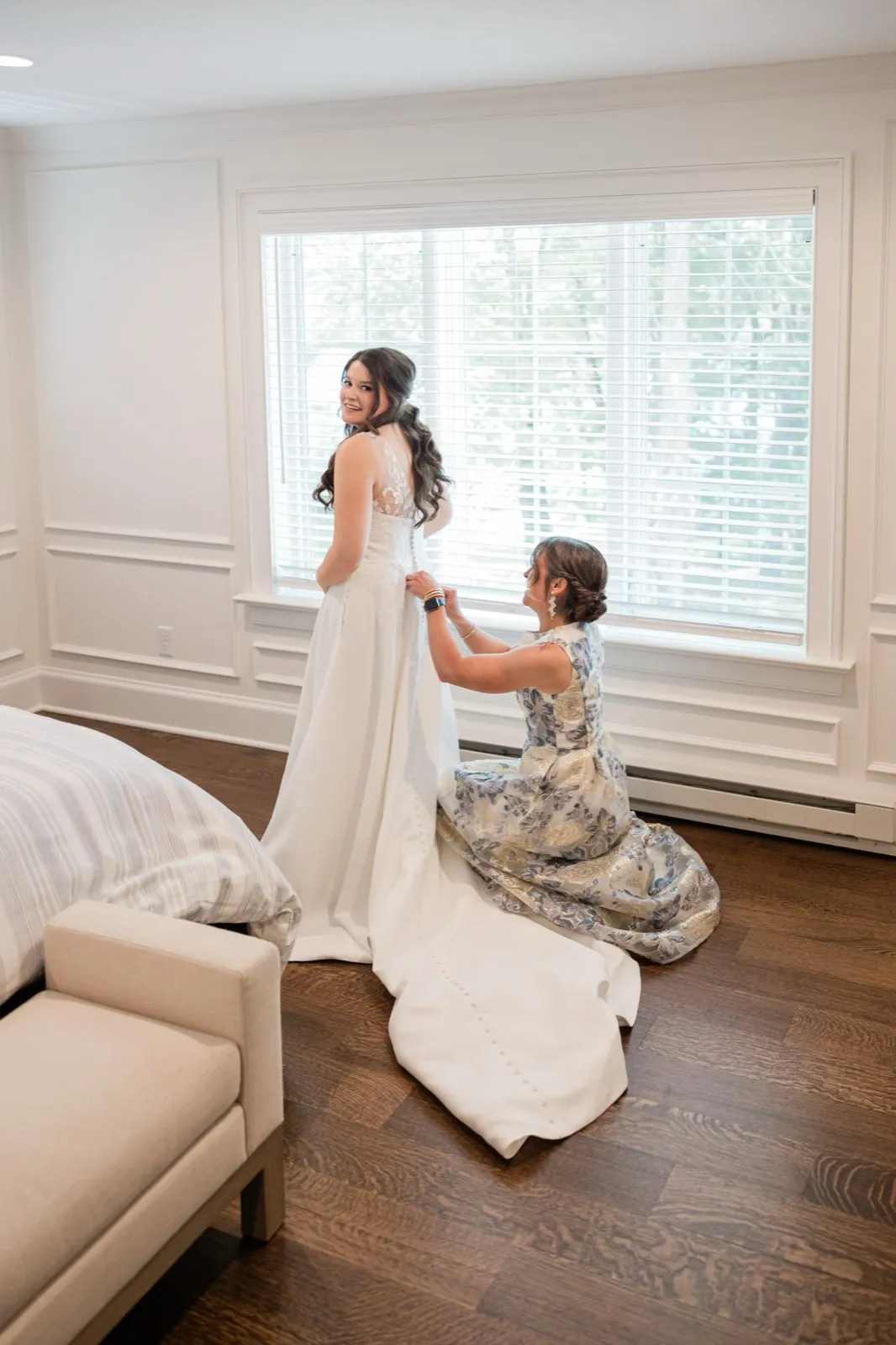 The bride stands in her full gown glancing back over her shoulder as her mother fastens it, the train pooled across the wood floor — Tim Larsen Photography, Brainerd Lakes MN