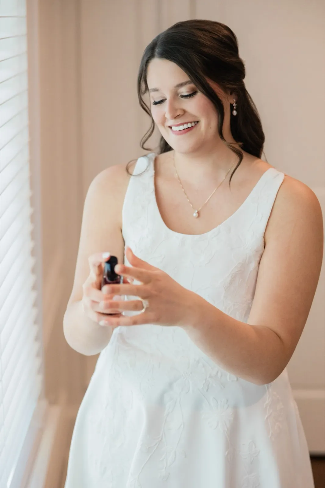The bride, dressed in her gown and pearl jewelry, smiles softly while spraying perfume on her wrist in warm window light — Tim Larsen Photography, Brainerd Lakes MN