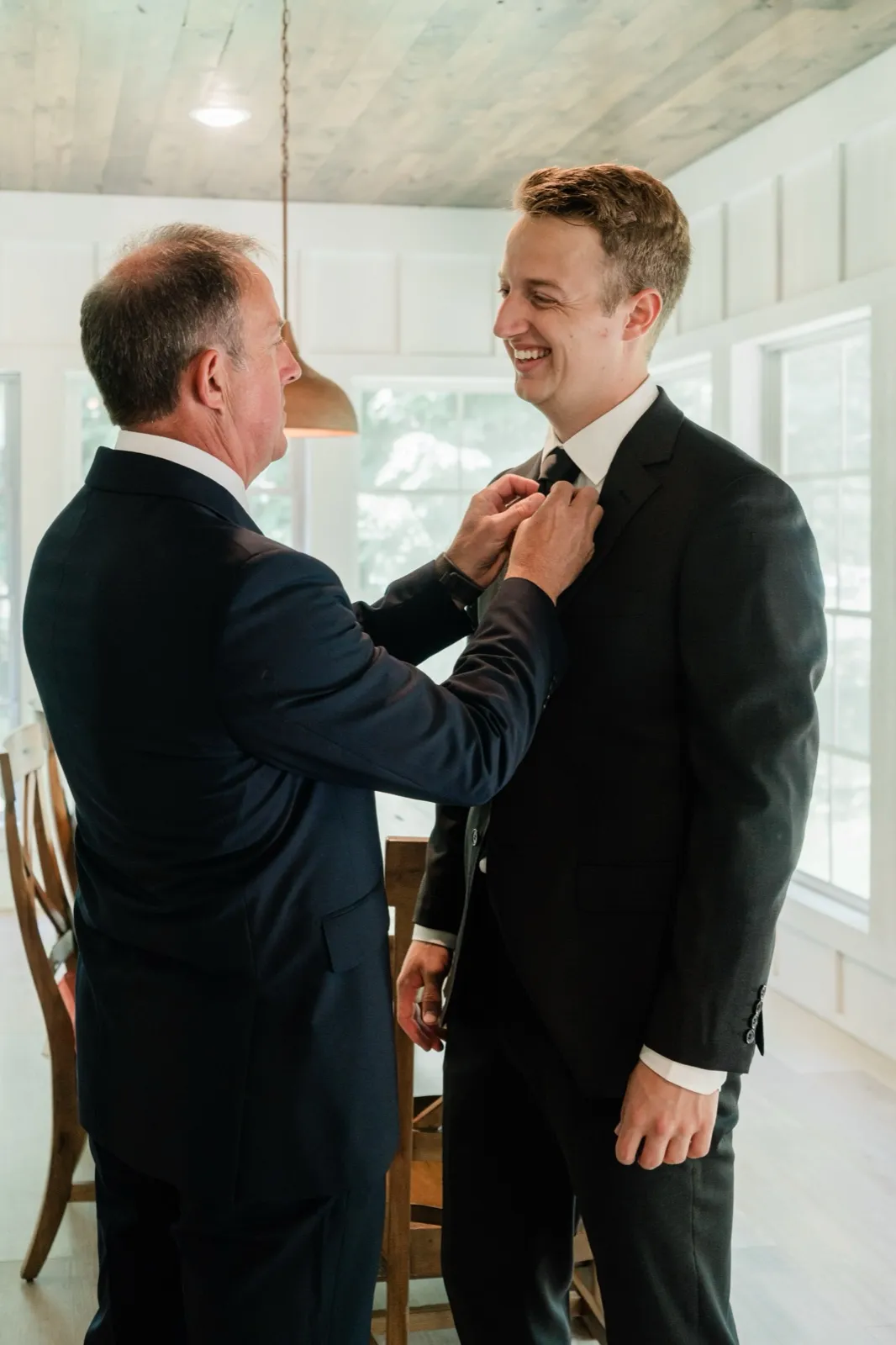 The groom grins as his father straightens his tie in the sunroom of a lakeside cabin — Tim Larsen Photography, Brainerd Lakes MN