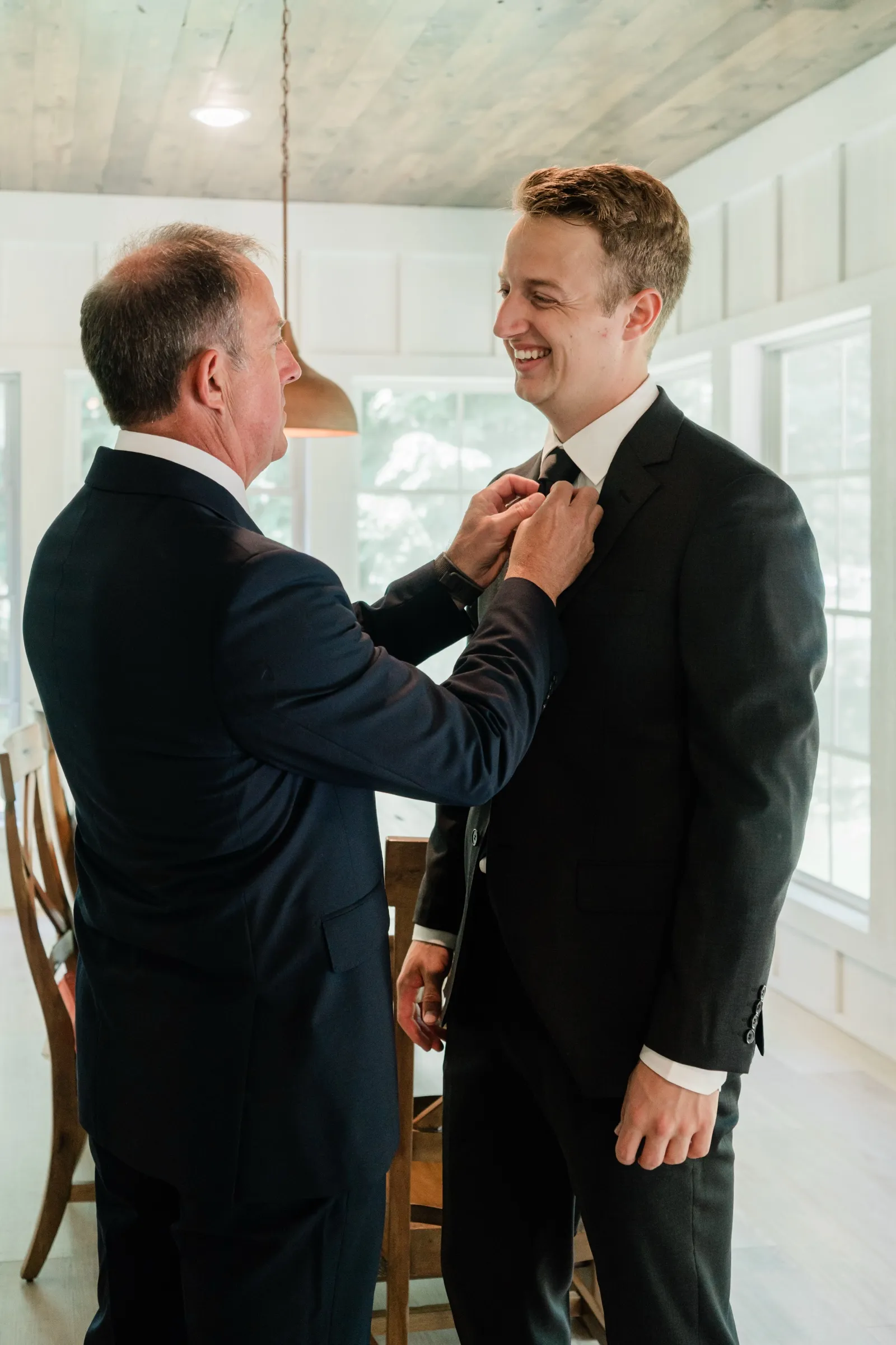 The groom grins as his father straightens his tie in the sunroom of a lakeside cabin — Tim Larsen Photography, Brainerd Lakes MN