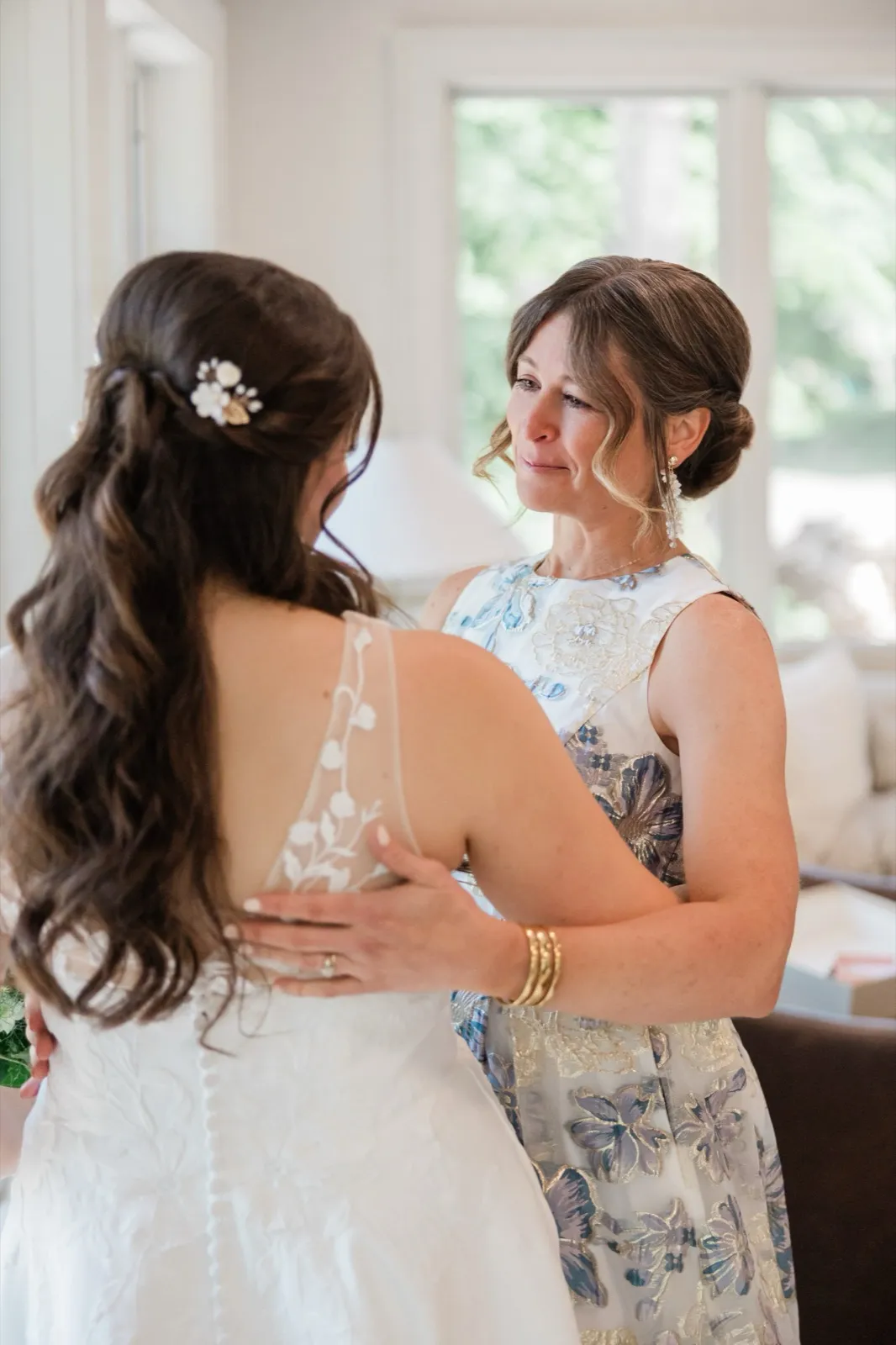 The bride's mother looks at her with welling eyes, hands resting on the bride's back near a sunlit window — Tim Larsen Photography, Brainerd Lakes MN