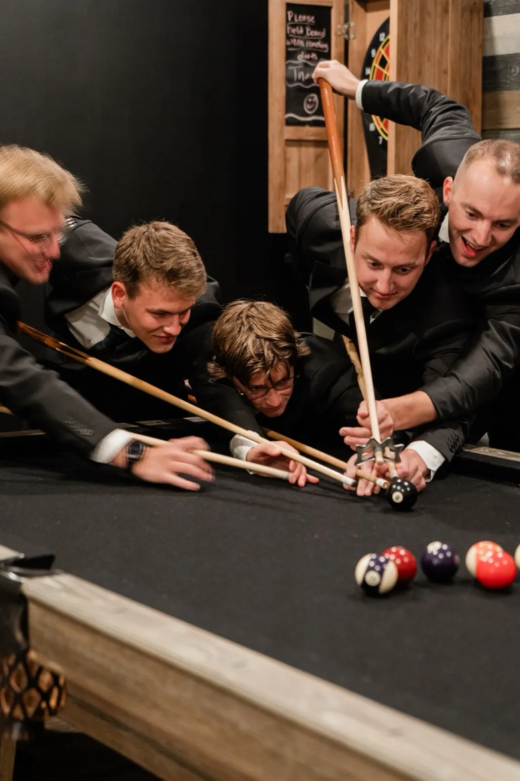 Five groomsmen crowd around a pool table, cue sticks converging on a single ball as they laugh together — Tim Larsen Photography, Brainerd Lakes MN
