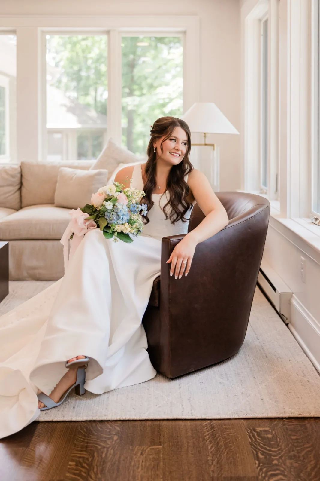 The bride sits in a leather armchair with her gown's train pooled on the floor and her bouquet in her lap, looking off toward the windows — Tim Larsen Photography, Brainerd Lakes MN