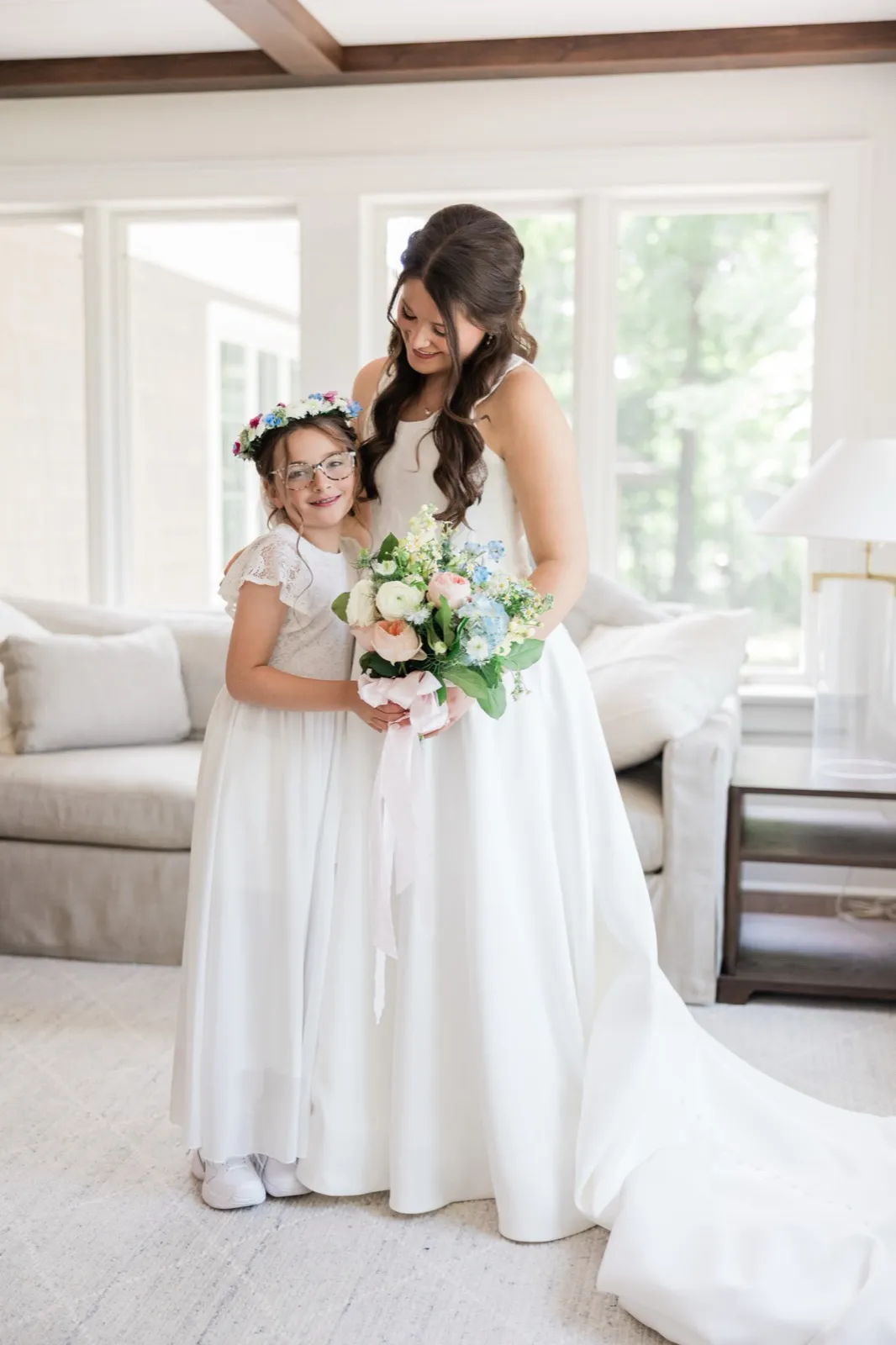 The bride gazes down at a young flower girl in a flower crown who holds the bride's bouquet, both in white in a sunlit room — Tim Larsen Photography, Brainerd Lakes MN