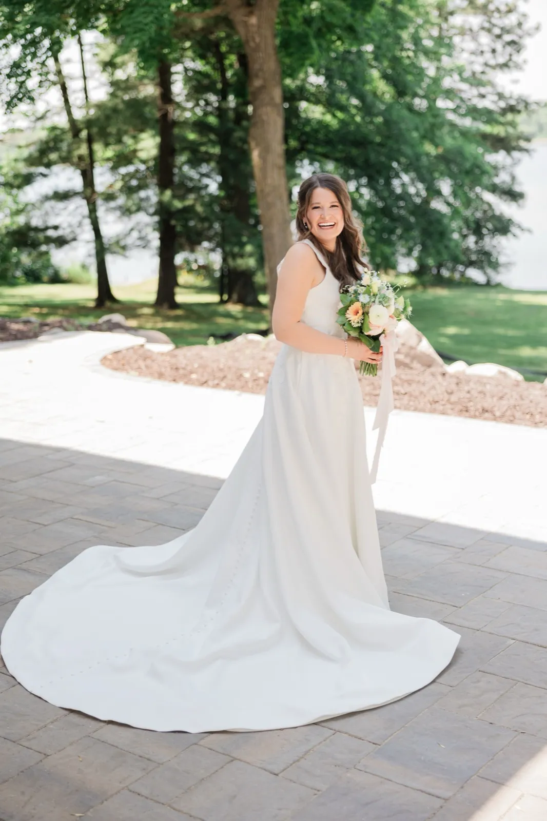 The bride laughs while standing on a paver patio, her gown's long train fanned out across the ground with trees and lake behind — Tim Larsen Photography, Brainerd Lakes MN