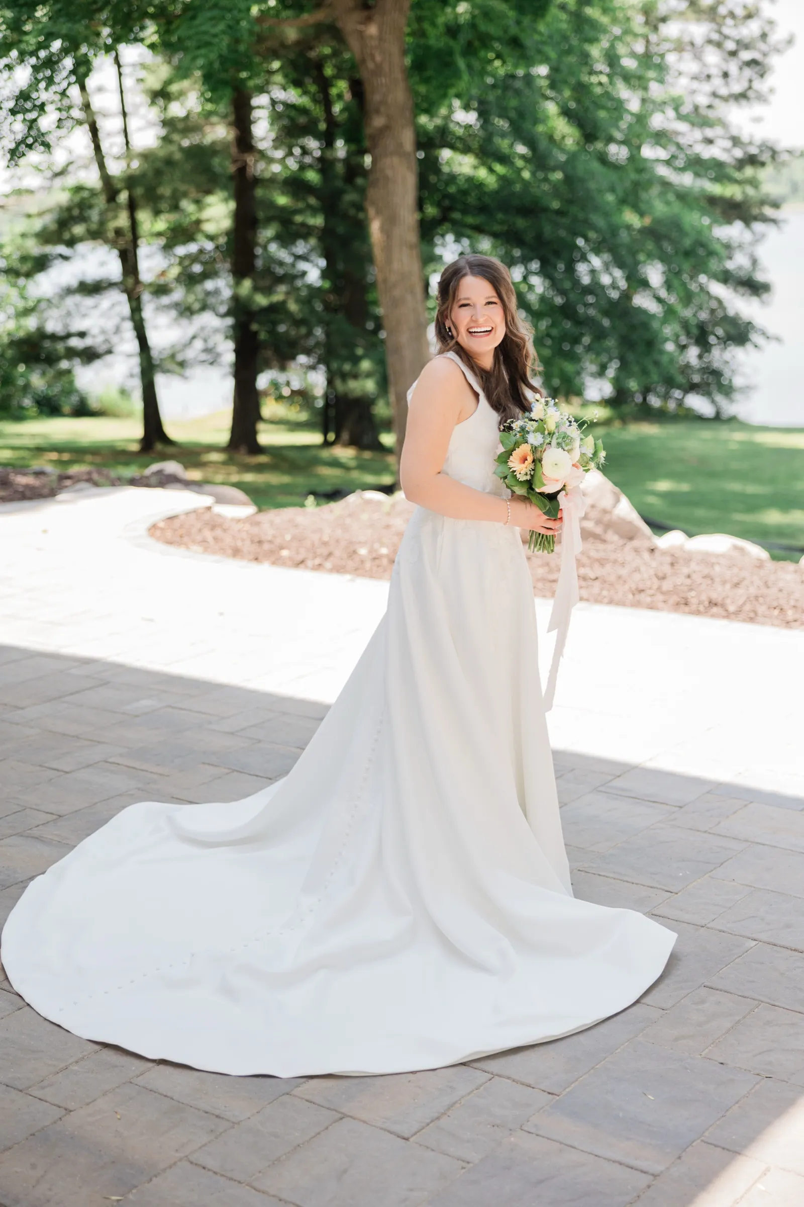 The bride laughs while standing on a paver patio, her gown's long train fanned out across the ground with trees and lake behind — Tim Larsen Photography, Brainerd Lakes MN