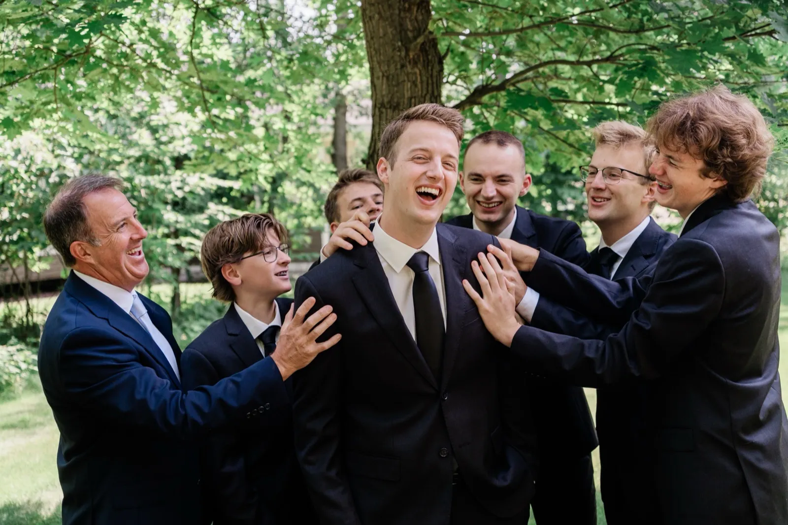 The groom laughs as his groomsmen and father crowd in around him, hands on his shoulders, beneath green trees — Tim Larsen Photography, Brainerd Lakes MN