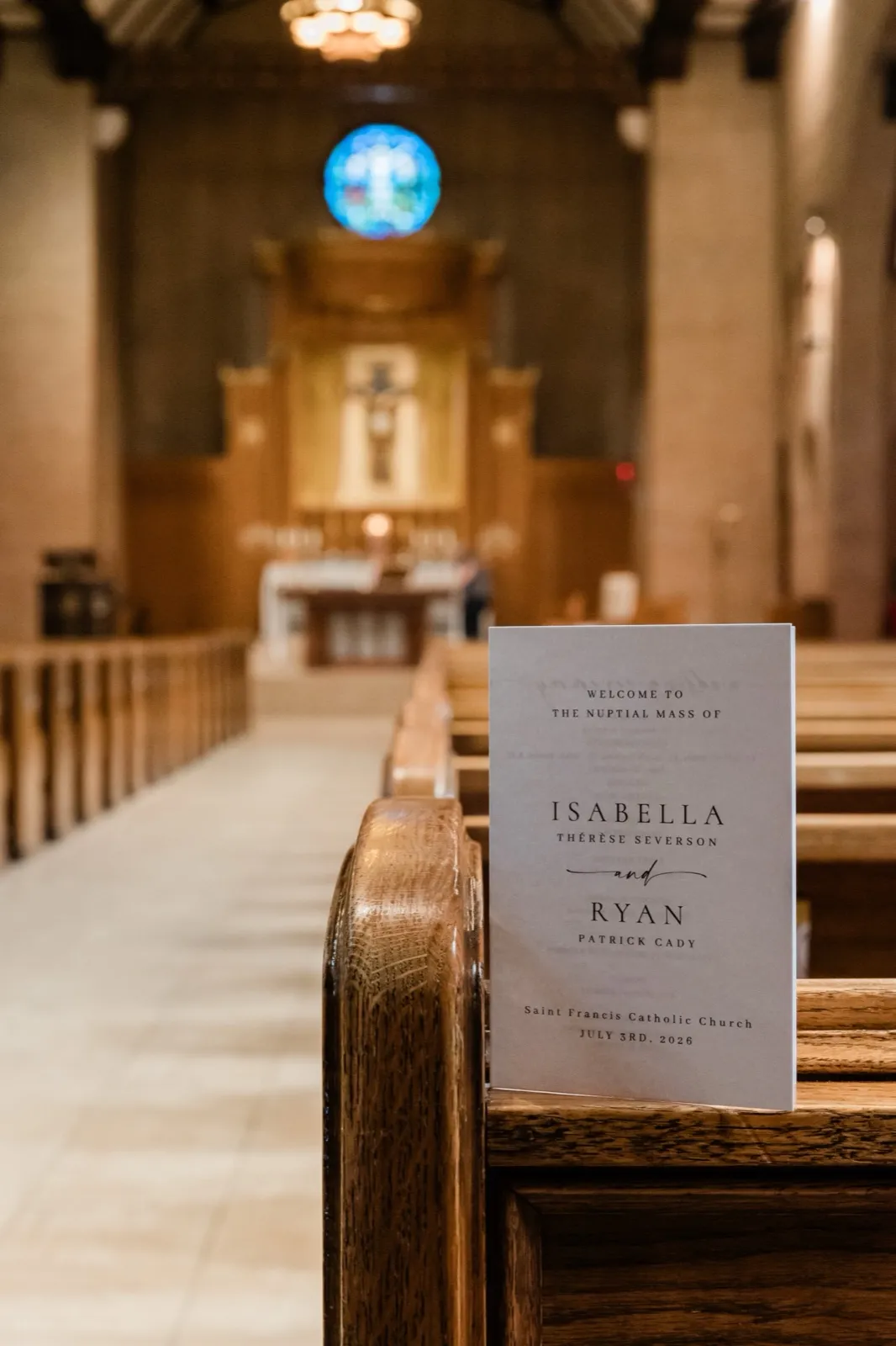 The nuptial Mass program for Isabella and Ryan rests on a pew rail inside St. Francis Catholic Church in Brainerd, the altar and round blue stained-glass window soft behind it — Tim Larsen Photography, Brainerd Lakes MN