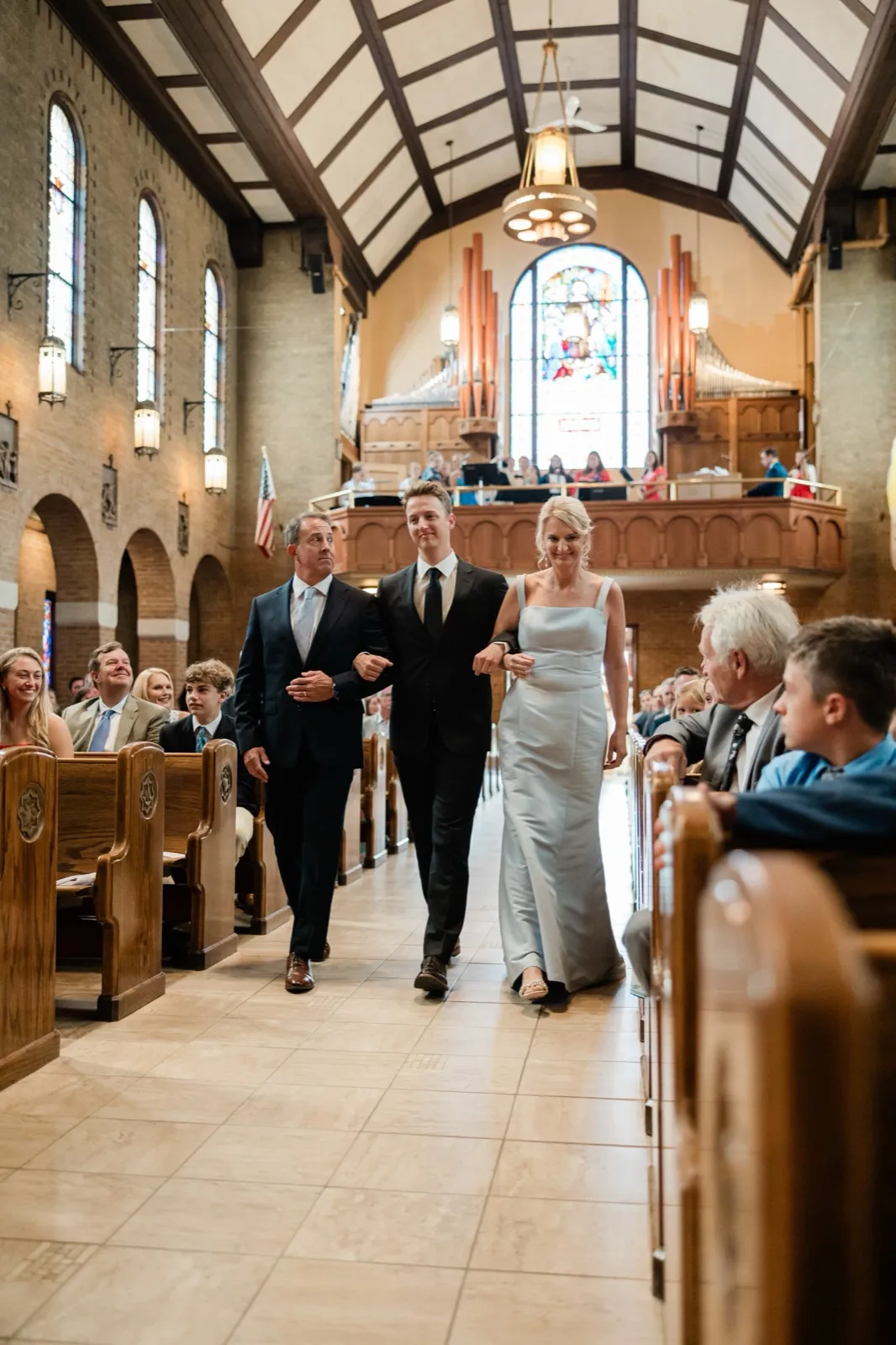 A groomsman walks up the aisle arm-in-arm with his parents through the grand nave of a Catholic church, guests seated in the pews beneath a soaring stained-glass window — Tim Larsen Photography, Brainerd Lakes MN