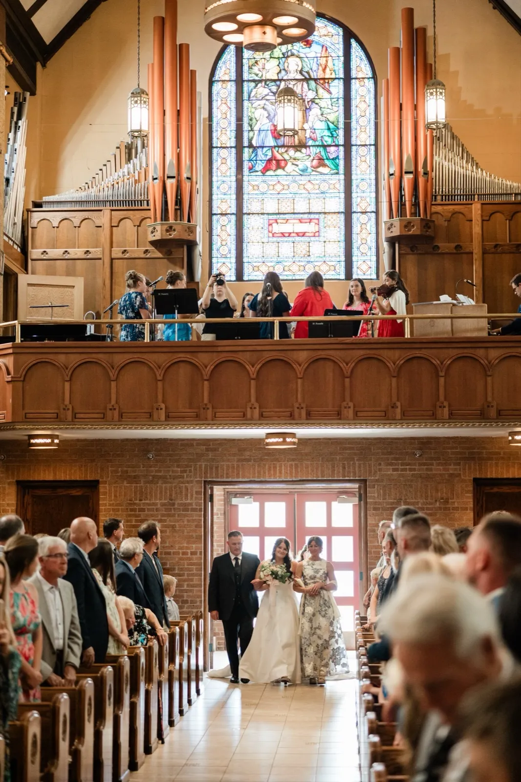 Seen from the front of the church, the bride stands at the far doorway with her parents as the standing congregation turns toward her beneath a stained-glass window and choir loft — Tim Larsen Photography, Brainerd Lakes MN