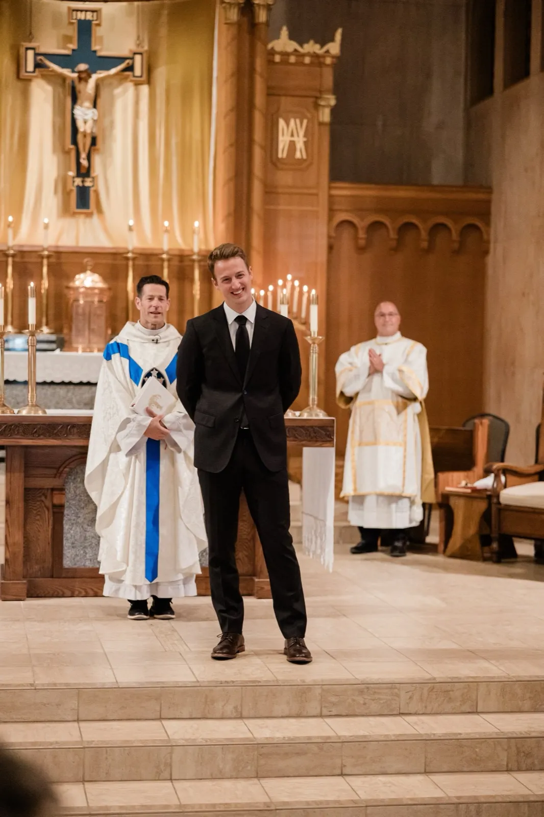The groom beams from the altar steps as his bride comes up the aisle at St. Francis Catholic Church in Brainerd, a priest and deacon behind him — Tim Larsen Photography, Brainerd Lakes MN