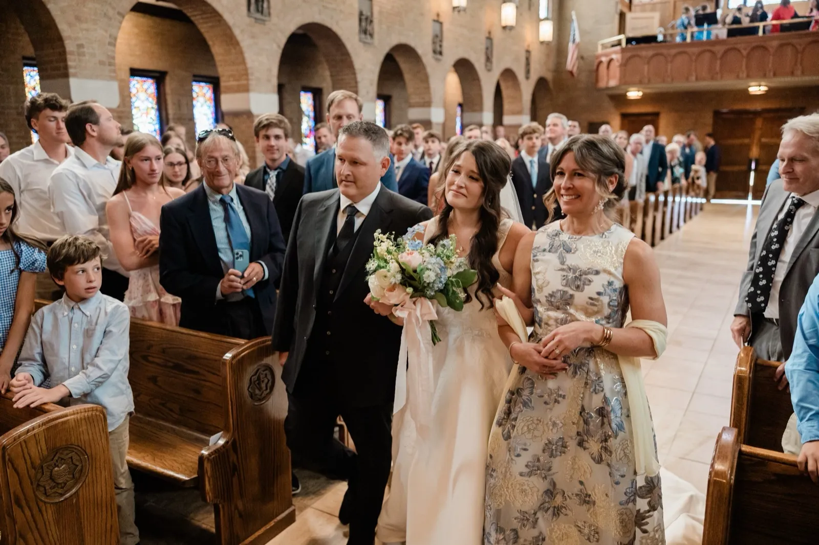 The bride walks up the aisle between her parents, her eyes filling with tears, as standing guests watch warmly from the pews — Tim Larsen Photography, Brainerd Lakes MN