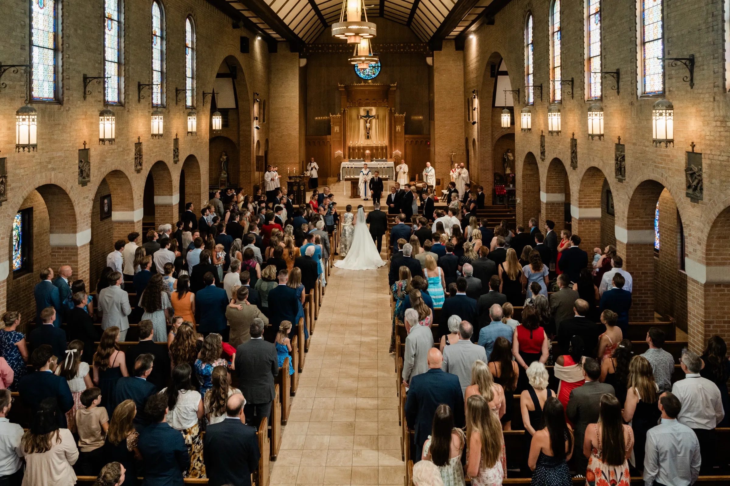 A wide view from the rear balcony looks down the full nave of a Catholic church, the standing congregation filling the pews as the couple stands at the distant altar — Tim Larsen Photography, Brainerd Lakes MN