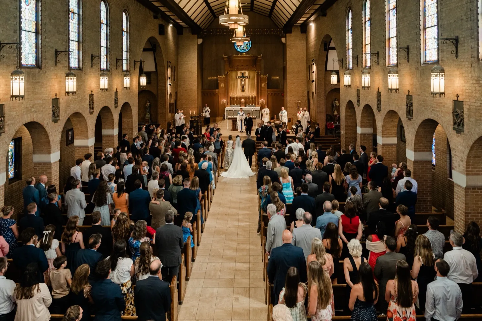 A wide view from the rear balcony looks down the full nave of a Catholic church, the standing congregation filling the pews as the couple stands at the distant altar — Tim Larsen Photography, Brainerd Lakes MN