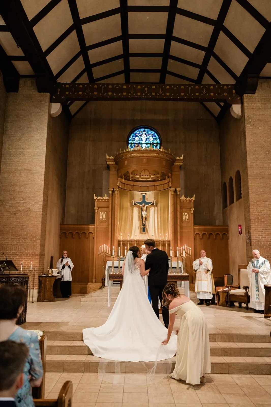The bride and groom stand together at the altar beneath a soaring vaulted ceiling and blue rose window, while a bridesmaid crouches to arrange the bride's long train — Tim Larsen Photography, Brainerd Lakes MN